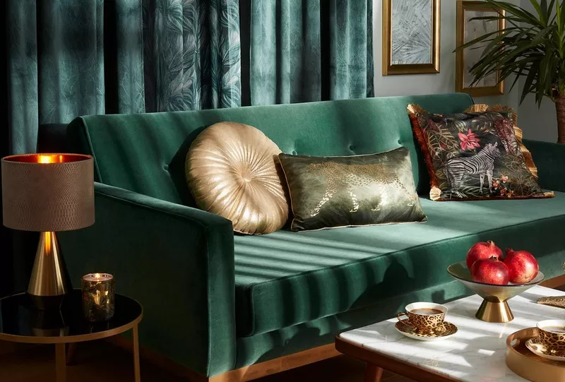 Elegancki zielony salon Pepco z aksamitną sofą, dekoracyjnymi poduszkami i lampą stołową, uzupełniony owocami granatu na stoliku.